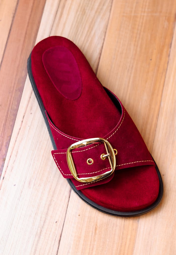 Elegant red suede slide sandal with a large gold buckle on a wooden floor background.